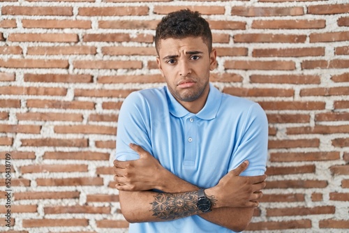 Brazilian young man standing over brick wall shaking and freezing for winter cold with sad and shock expression on face