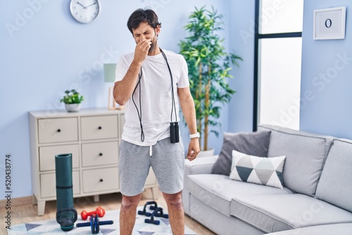 Handsome latin man wearing sportswear at home smelling something stinky and disgusting, intolerable smell, holding breath with fingers on nose. bad smell