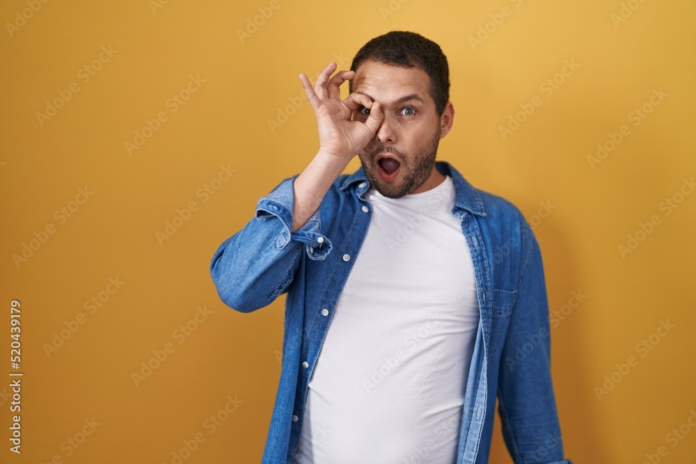 Hispanic man standing over yellow background doing ok gesture shocked with surprised face, eye looking through fingers. unbelieving expression. Hispanic man standing over yellow background doing ok gesture shocked with surprised face, eye looking through fingers. unbelieving expression.