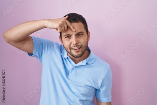 Hispanic man standing over pink background pointing unhappy to pimple on forehead, ugly infection of blackhead. acne and skin problem