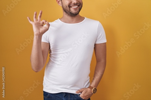 Hispanic man wearing white t shirt over yellow background doing ok sign with fingers, smiling friendly gesturing excellent symbol