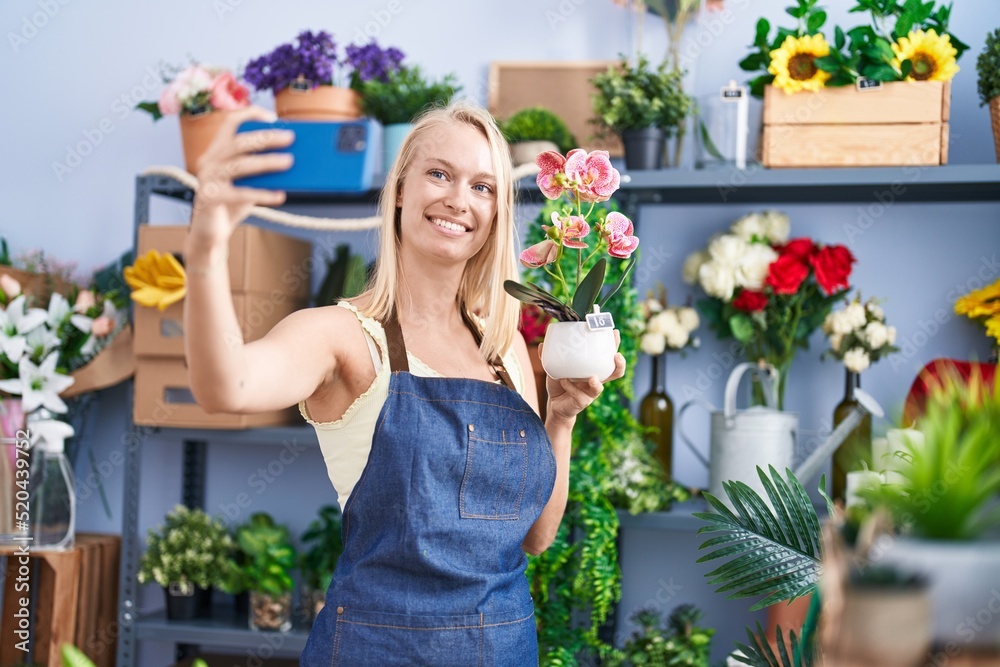 Young blonde woman florist make selfie by smartphone holding plant at florist store