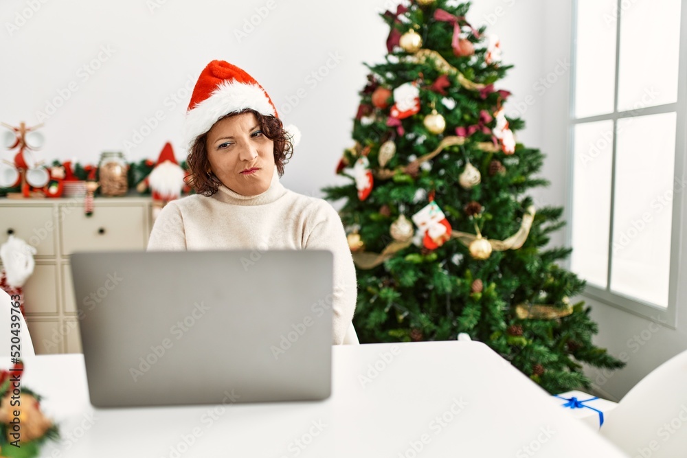 Middle age hispanic woman using laptop sitting by christmas tree skeptic and nervous, disapproving expression on face with crossed arms. negative person. Middle age hispanic woman using laptop sitting by christmas tree skeptic and nervous, disapproving expression on face with crossed arms. negative person.