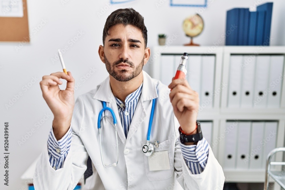 Young doctor man holding electronic cigarette at medical clinic relaxed with serious expression on face. simple and natural looking at the camera. Young doctor man holding electronic cigarette at medical clinic relaxed with serious expression on face. simple and natural looking at the camera.