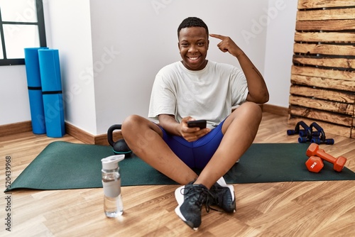 Young african man sitting on training mat at the gym using smartphone smiling pointing to head with one finger, great idea or thought, good memory