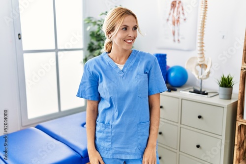 Beautiful blonde physiotherapist woman working at pain recovery clinic looking away to side with smile on face  natural expression. laughing confident.