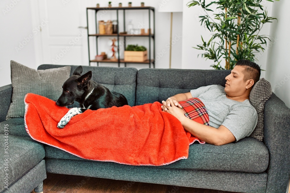 Young hispanic man sleeping covering with blanket lying on the sofa with dog at home.