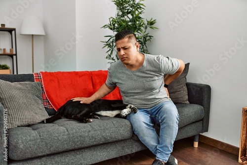 Young latin man and dog sitting on the sofa at home suffering of backache, touching back with hand, muscular pain