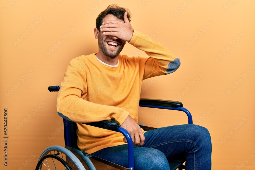 Handsome man with beard sitting on wheelchair smiling and laughing with hand on face covering eyes for surprise. blind concept.