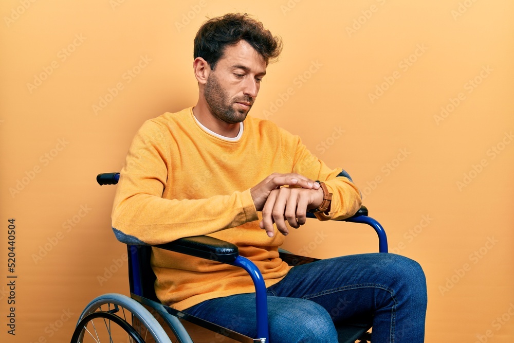 Handsome man with beard sitting on wheelchair checking the time on wrist watch, relaxed and confident Handsome man with beard sitting on wheelchair checking the time on wrist watch, relaxed and confident