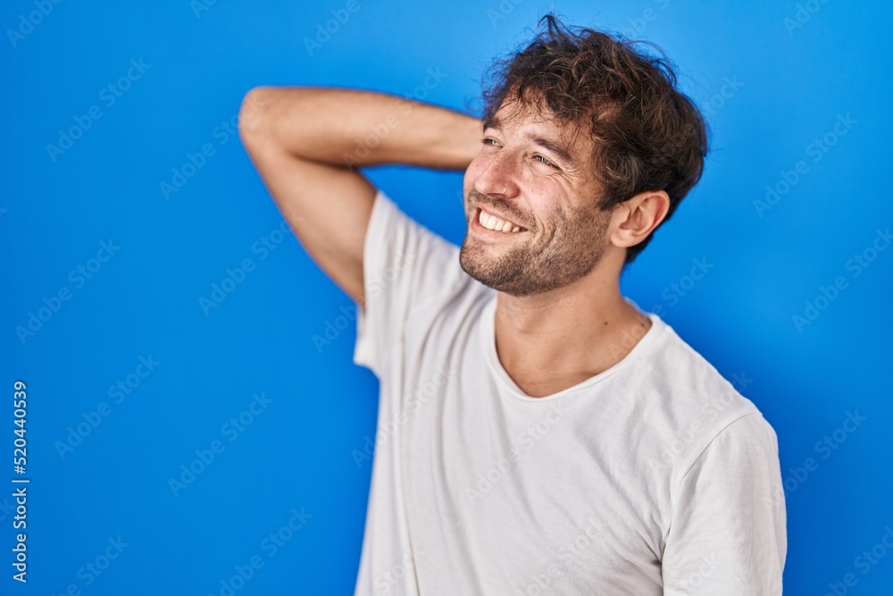 Hispanic young man standing over blue background smiling confident touching hair with hand up gesture, posing attractive and fashionable Hispanic young man standing over blue background smiling confident touching hair with hand up gesture, posing attractive and fashionable