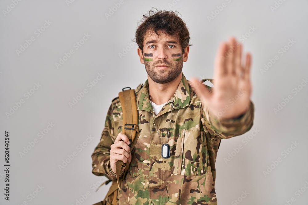 Hispanic young man wearing camouflage army uniform doing stop sing with palm of the hand. warning expression with negative and serious gesture on the face. Hispanic young man wearing camouflage army uniform doing stop sing with palm of the hand. warning expression with negative and serious gesture on the face.