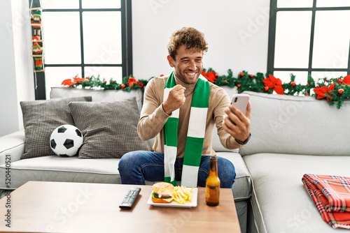 Fototapeta Naklejka Na Ścianę i Meble -  Young hispanic man supporting soccer match using smartphone sitting by christmas decor at home