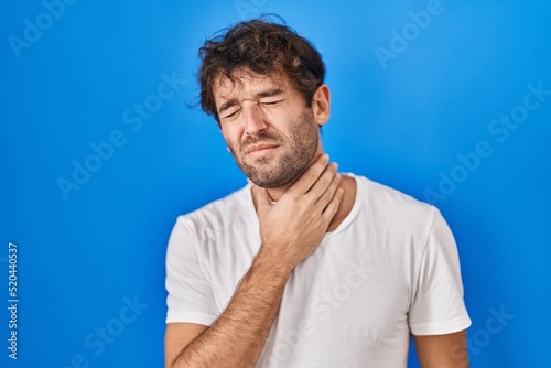 Hispanic young man standing over blue background touching painful neck, sore throat for flu, clod and infection