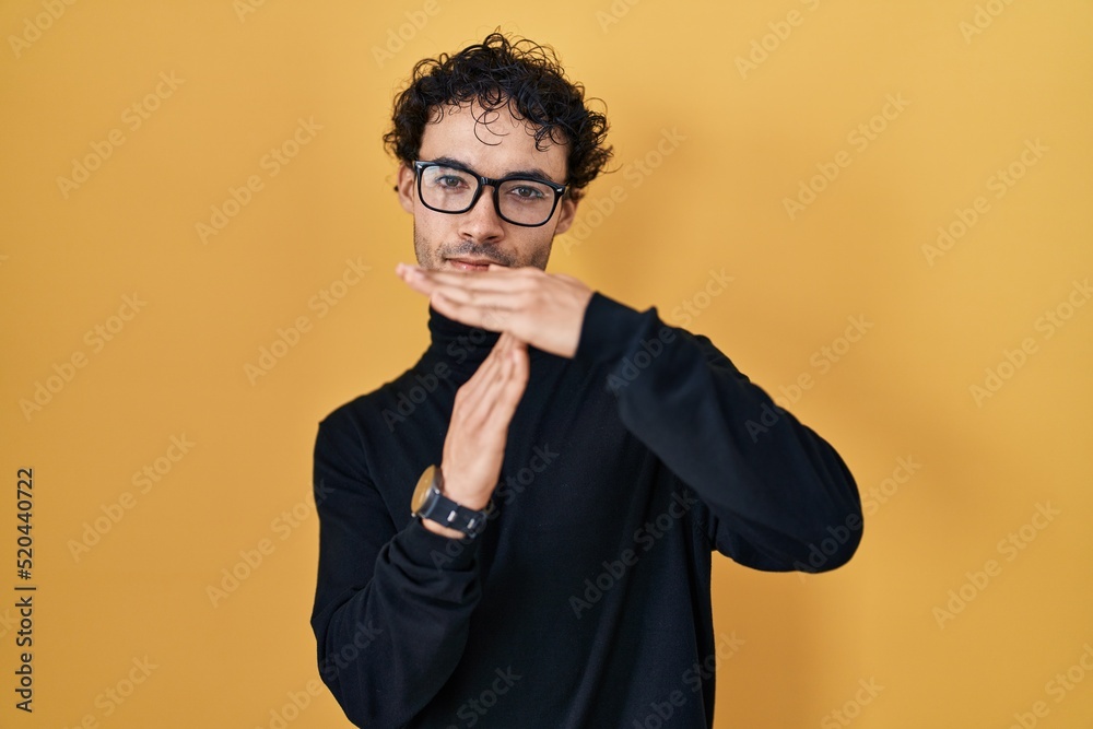 Hispanic man standing over yellow background doing time out gesture with hands, frustrated and serious face