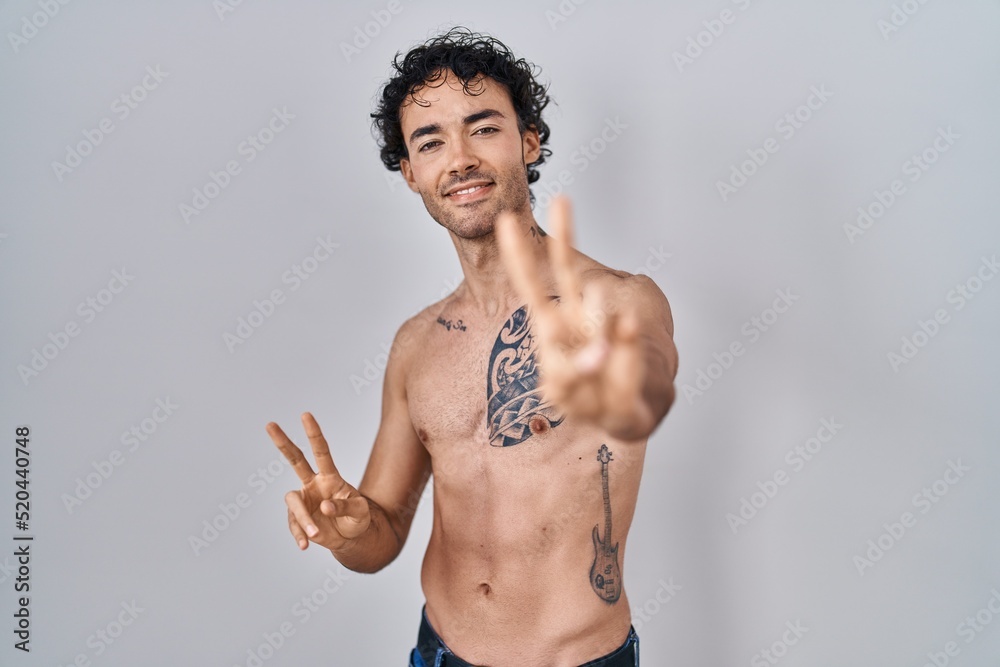 Fototapeta premium Hispanic man standing shirtless smiling looking to the camera showing fingers doing victory sign. number two.