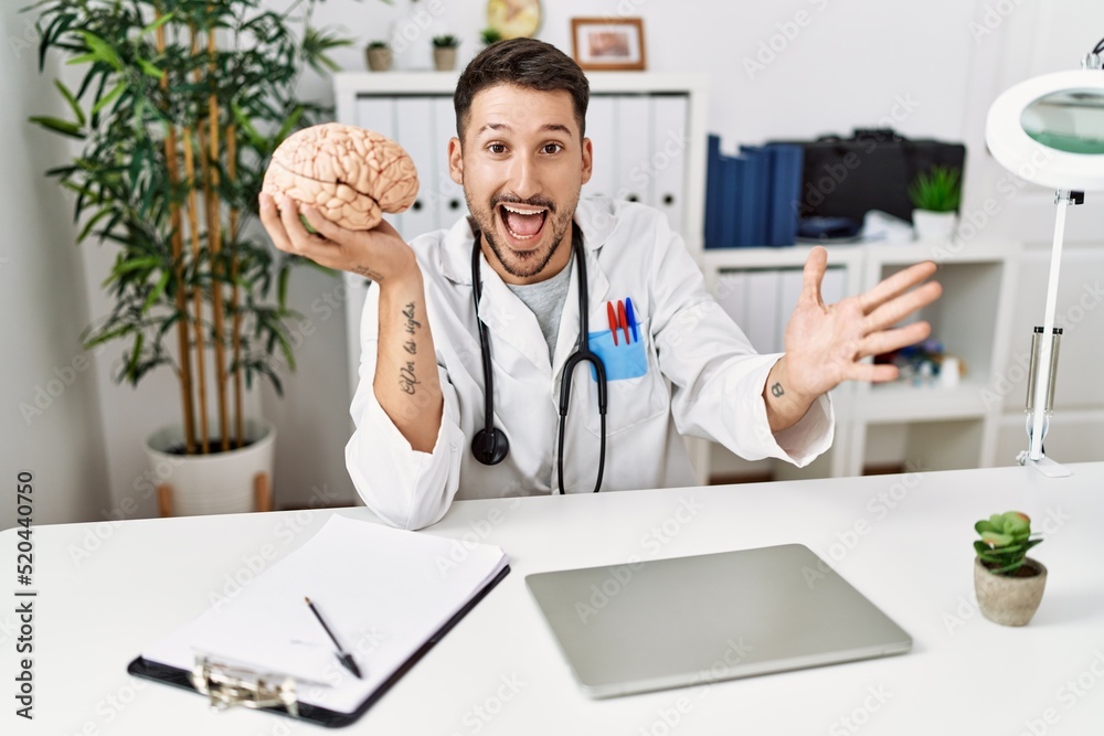 Obraz premium Young doctor holding brain at medical clinic celebrating victory with happy smile and winner expression with raised hands