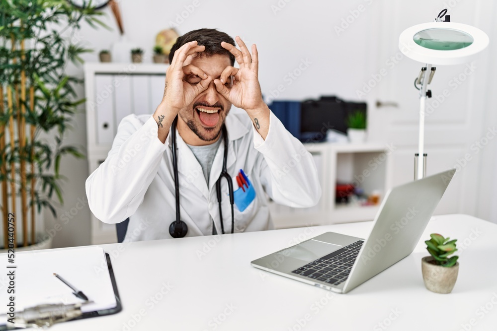 Young doctor working at the clinic using computer laptop doing ok gesture like binoculars sticking tongue out, eyes looking through fingers. crazy expression.