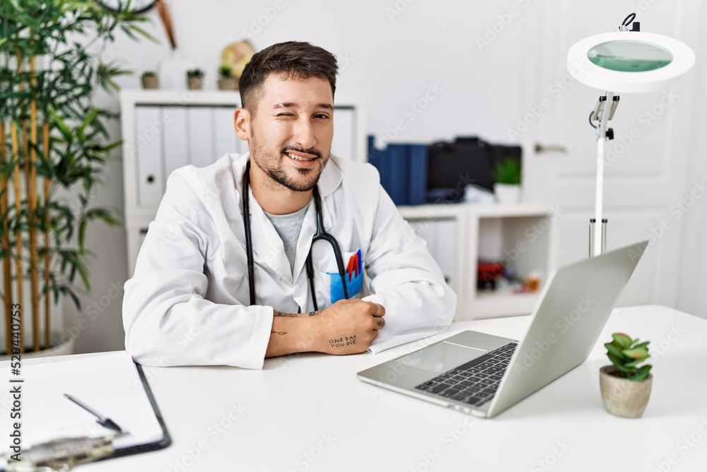 Young doctor working at the clinic using computer laptop winking looking at the camera with sexy expression, cheerful and happy face. Young doctor working at the clinic using computer laptop winking looking at the camera with sexy expression, cheerful and happy face.