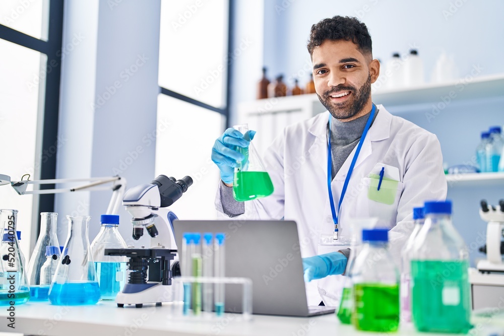Young hispanic man wearing scientist uniform using laptop holding test tube at laboratory Young hispanic man wearing scientist uniform using laptop holding test tube at laboratory