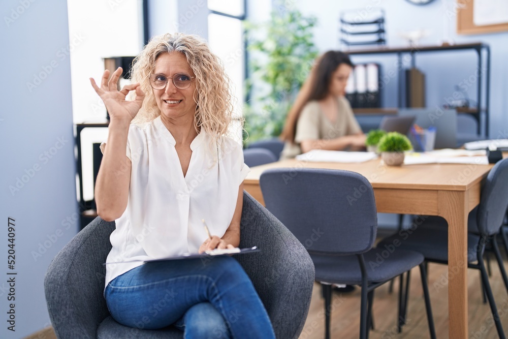 Middle age woman working at the office holding clipboard doing ok sign with fingers, smiling friendly gesturing excellent symbol Middle age woman working at the office holding clipboard doing ok sign with fingers, smiling friendly gesturing excellent symbol