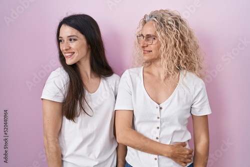 Mother and daughter standing together over pink background looking away to side with smile on face, natural expression. laughing confident.