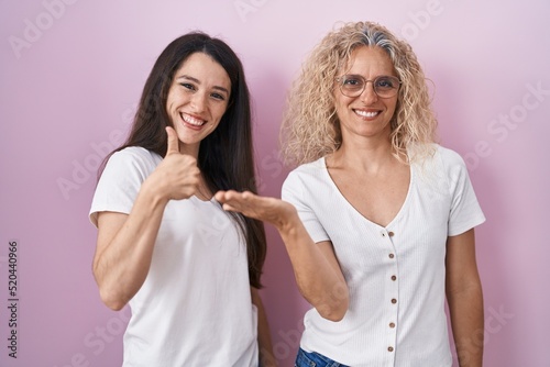 Mother and daughter standing together over pink background showing palm hand and doing ok gesture with thumbs up, smiling happy and cheerful