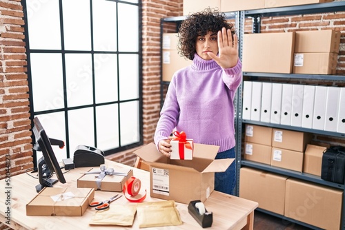 Young brunette woman with curly hair working at small business ecommerce preparing order with open hand doing stop sign with serious and confident expression, defense gesture