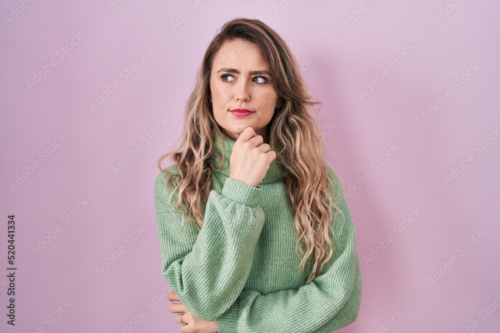 Young caucasian woman standing over pink background looking confident at the camera with smile with crossed arms and hand raised on chin. thinking positive.