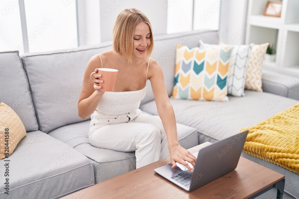 Young blonde woman using laptop and drinking coffee sitting on sofa at home Young blonde woman using laptop and drinking coffee sitting on sofa at home