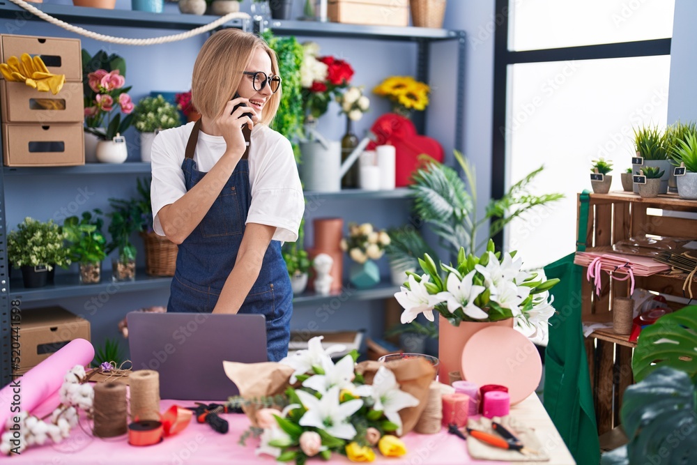 Young blonde woman florist using laptop talking on smartphone at florist shop