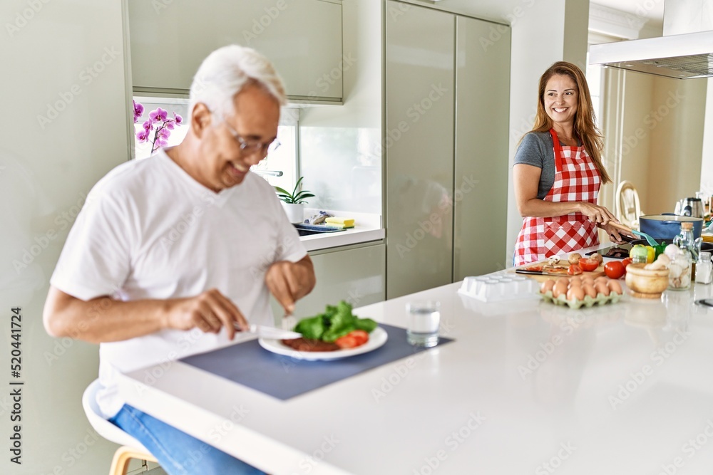 Middle age hispanic man smiling happy eating beef with salad while woman cook at the kitchen.