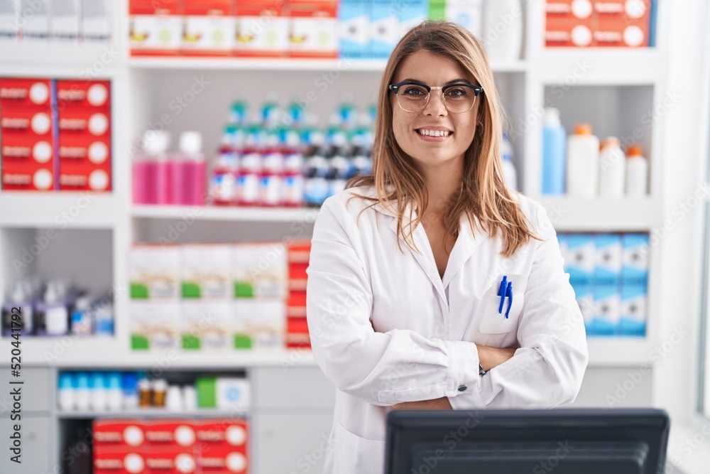 Young woman pharmacist smiling confident standing with arms crossed gesture at pharmacy