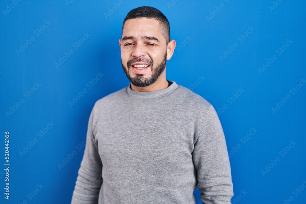Hispanic man standing over blue background winking looking at the camera with sexy expression, cheerful and happy face. Hispanic man standing over blue background winking looking at the camera with sexy expression, cheerful and happy face.