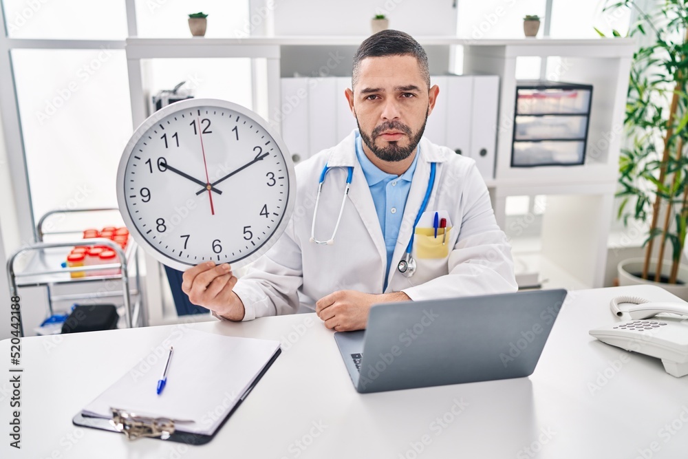 Hispanic doctor man holding clock at the clinic thinking attitude and sober expression looking self confident