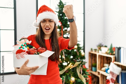 Young brunette woman standing by christmas tree holding decoration angry and mad raising fist frustrated and furious while shouting with anger. rage and aggressive concept.