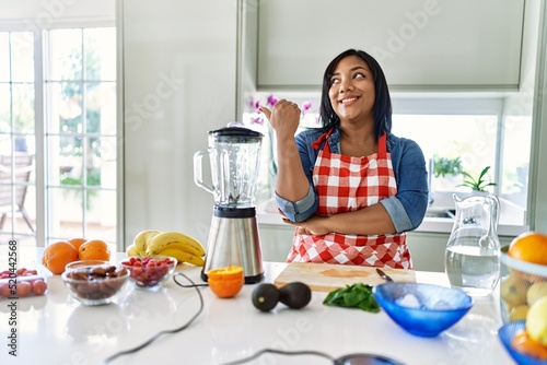 Young hispanic woman making healthy smoothie smiling with happy face looking and pointing to the side with thumb up.