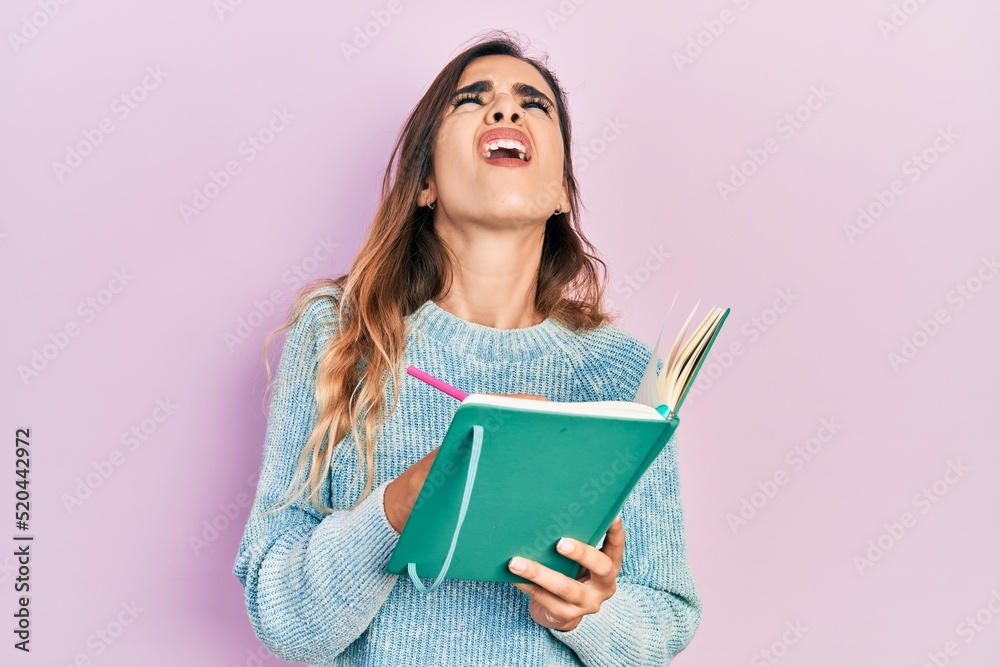 Young hispanic girl reading and writing book angry and mad screaming frustrated and furious, shouting with anger looking up. Young hispanic girl reading and writing book angry and mad screaming frustrated and furious, shouting with anger looking up.