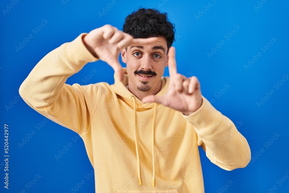 Fototapeta premium Hispanic man standing over blue background smiling making frame with hands and fingers with happy face. creativity and photography concept.