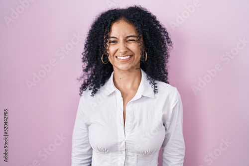 Hispanic woman with curly hair standing over pink background winking looking at the camera with sexy expression, cheerful and happy face.