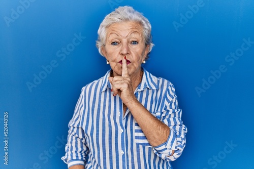 Senior woman with grey hair standing over blue background asking to be quiet with finger on lips. silence and secret concept.