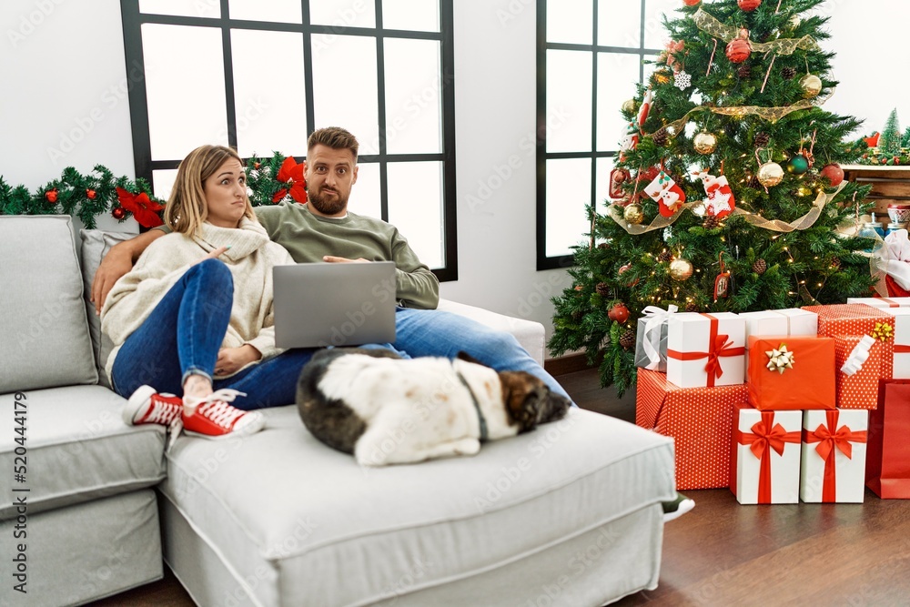 Young couple using laptop sitting by christmas tree pointing aside worried and nervous with forefinger, concerned and surprised expression Young couple using laptop sitting by christmas tree pointing aside worried and nervous with forefinger, concerned and surprised expression