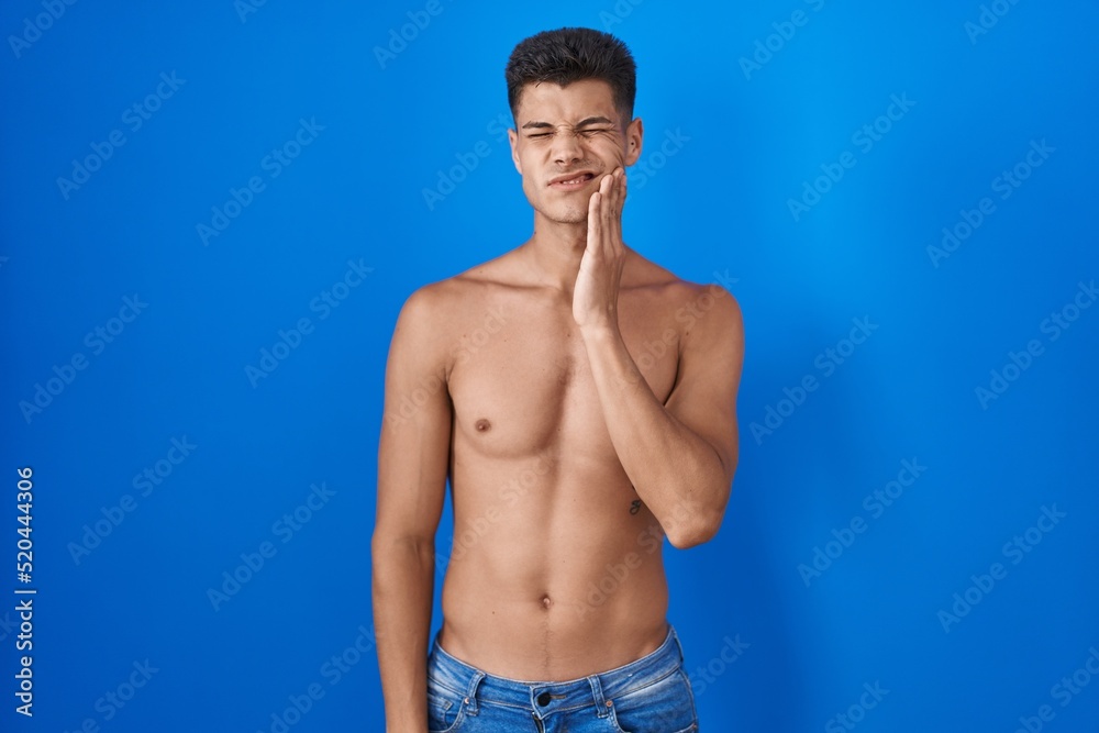 Young hispanic man standing shirtless over blue background touching mouth with hand with painful expression because of toothache or dental illness on teeth. dentist