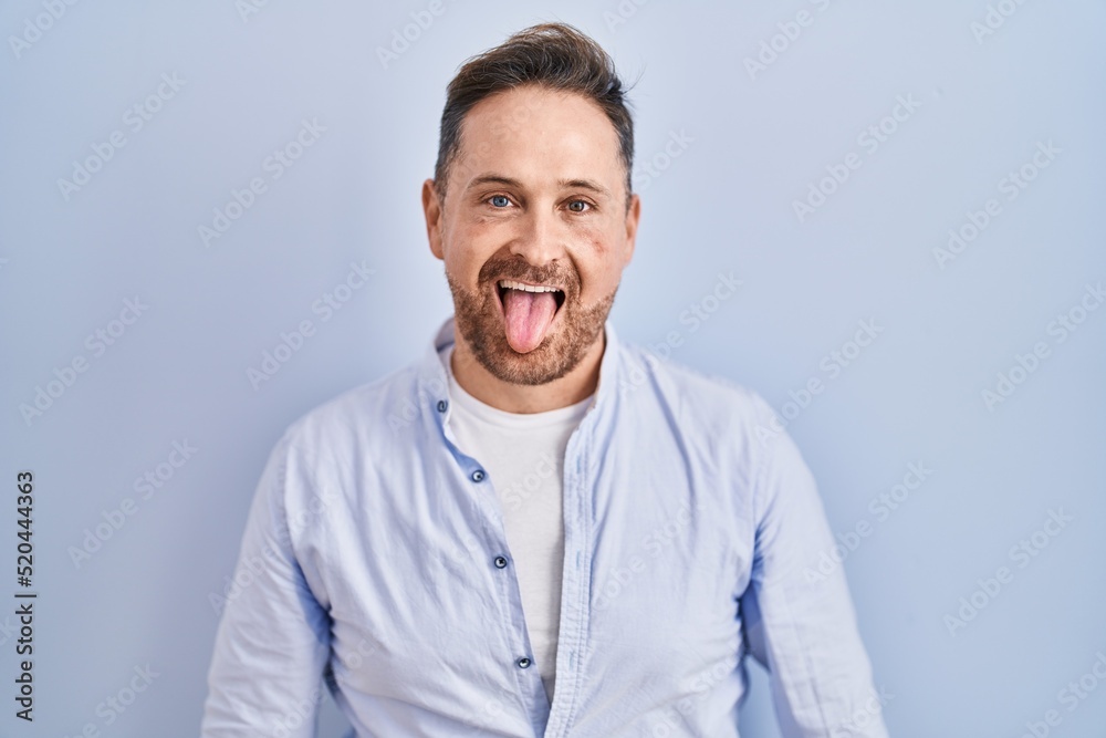 Middle age caucasian man standing over blue background sticking tongue out happy with funny expression. emotion concept.