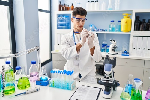 Young hispanic man wearing scientist uniform writing on test tube at laboratory