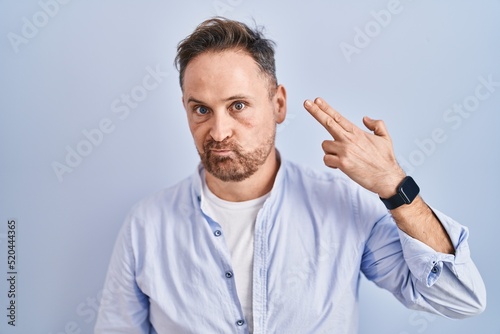 Middle age caucasian man standing over blue background shooting and killing oneself pointing hand and fingers to head like gun  suicide gesture.