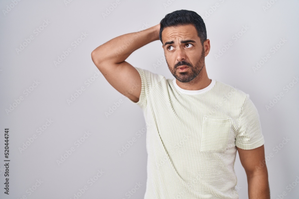 Hispanic man with beard standing over isolated background confuse and wondering about question. uncertain with doubt, thinking with hand on head. pensive concept. Hispanic man with beard standing over isolated background confuse and wondering about question. uncertain with doubt, thinking with hand on head. pensive concept.