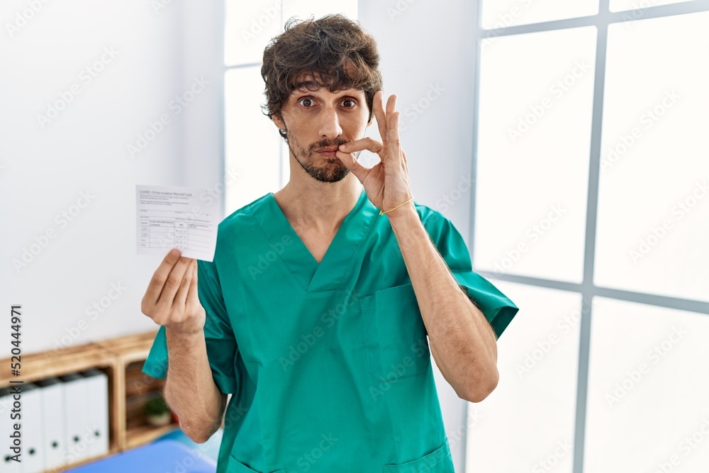 Foto de Young hispanic doctor man holding covid record card mouth and