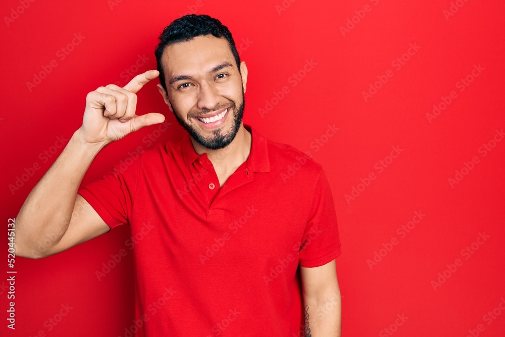 Hispanic man with beard wearing casual red t shirt smiling and confident gesturing with hand doing small size sign with fingers looking and the camera. measure concept. Hispanic man with beard wearing casual red t shirt smiling and confident gesturing with hand doing small size sign with fingers looking and the camera. measure concept.