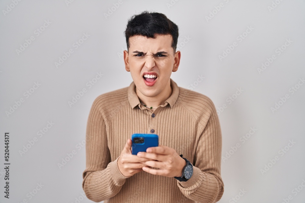 Non binary person using smartphone typing message angry and mad screaming frustrated and furious, shouting with anger. rage and aggressive concept. Non binary person using smartphone typing message angry and mad screaming frustrated and furious, shouting with anger. rage and aggressive concept.